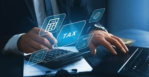 A graphic showing a professional working at a laptop with a calculator and the word Tax floating above his hands